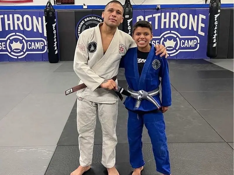 A coach and a young student in gis stand on the mats in front of the Dethrone Base Camp wall.