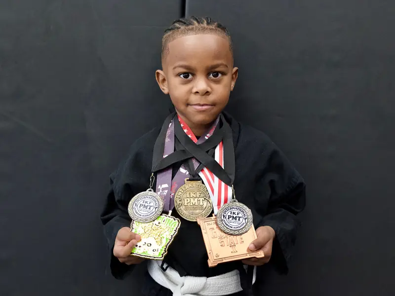 A young student in a gi holds up his tournament medals.