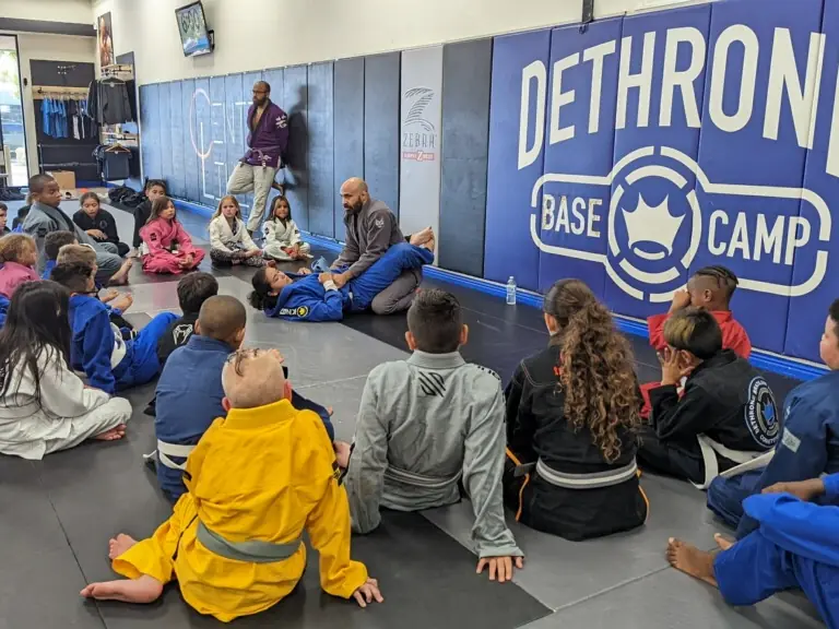 A kids class sits on the mats in front of the Dethrone Base Camp wall, listening to the coach.
