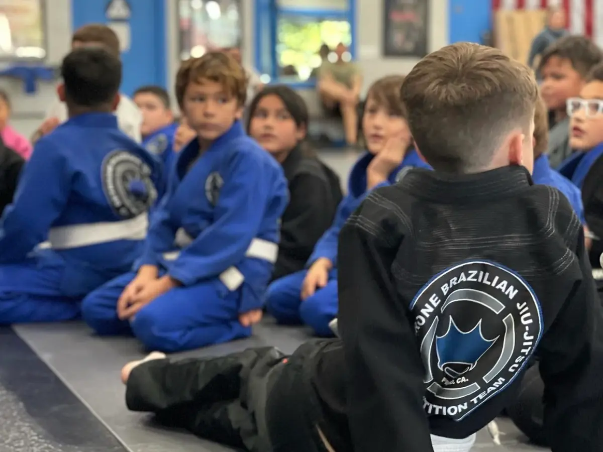 Kids in blue and black gis sit on the mats during a class.