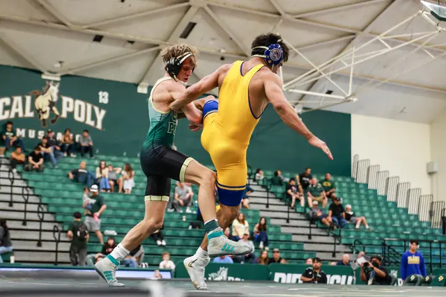 Two wrestlers, one in green and one in yellow, leave the mat in a scramble in the Cal Poly gym.