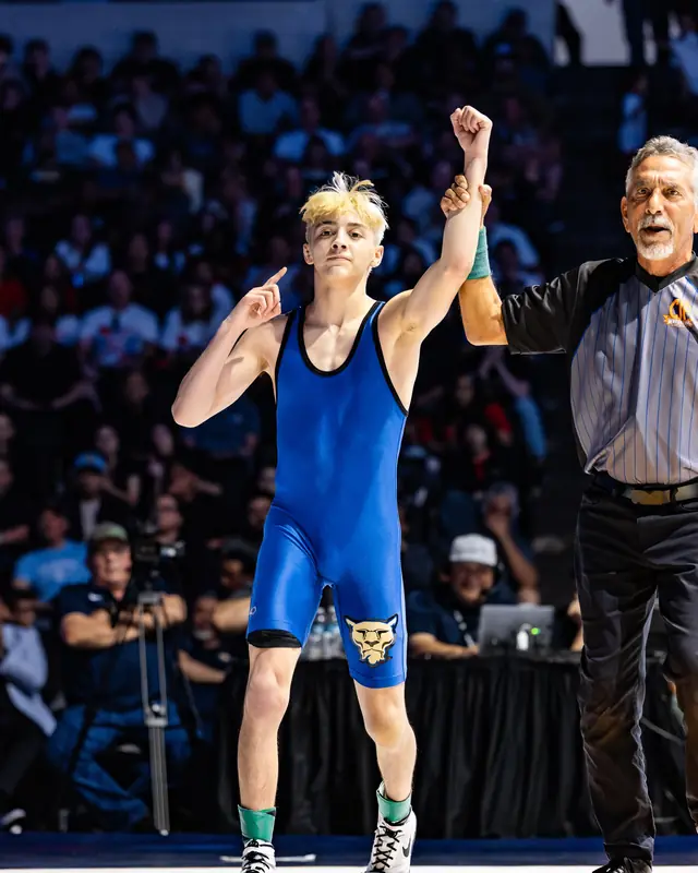 A referee raises the hand of Anthony Garza, in a blue singlet, on the mat.