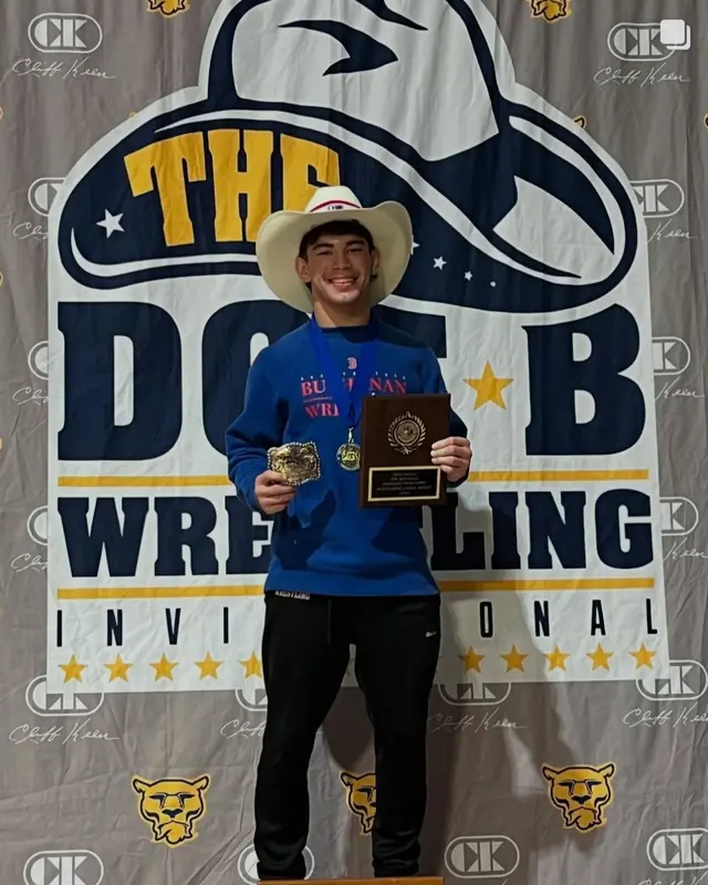Ashton Besmer in a cowboy hat, holding a medal and a plaque in front of the Doc B Wrestling Invitational banner.