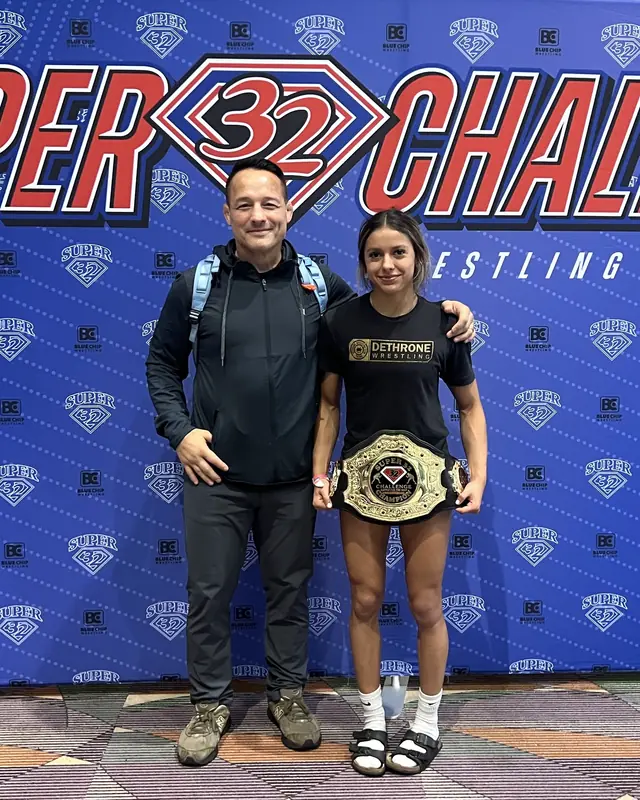 Christina Estrada, in a Dethrone Wrestling shirt, holds a championship belt in front of the Super 32 Challenge backdrop.