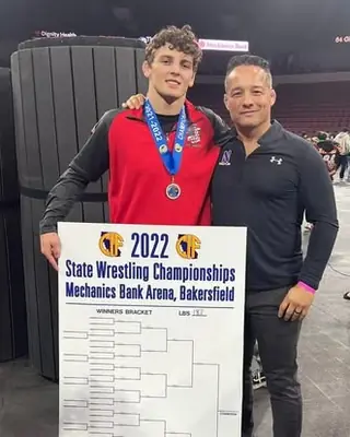 Joseph Martin, wearing a state medal, stands with Coach Jason Kraft beside the 2022 California state championships bracket in Bakersfield.