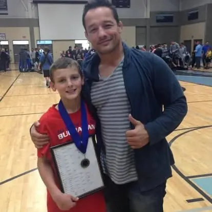 A young Joseph Martin, in a red Buchanan shirt, wears a medal and holds a plaque beside Coach Jason Kraft, who gives a thumbs-up.