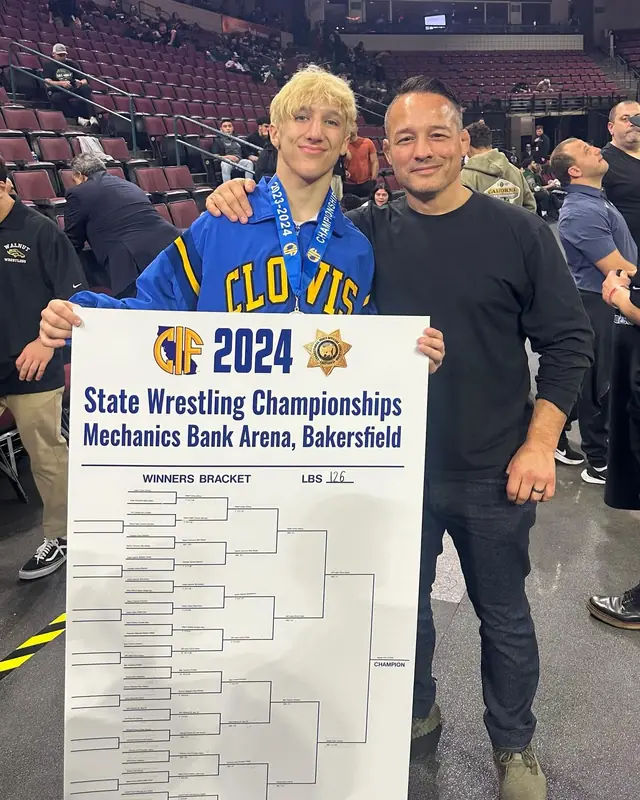 Nikade Zinkin, in a Clovis jacket and a medal, holds the 126-pound bracket from the 2024 CIF State Wrestling Championships in Bakersfield, beside Coach Jason Kraft.