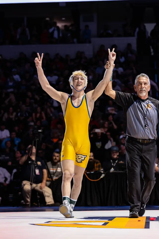 Nikade Zinkin, in a gold singlet, holds up a V sign with each hand as a referee raises his arm in a packed arena.