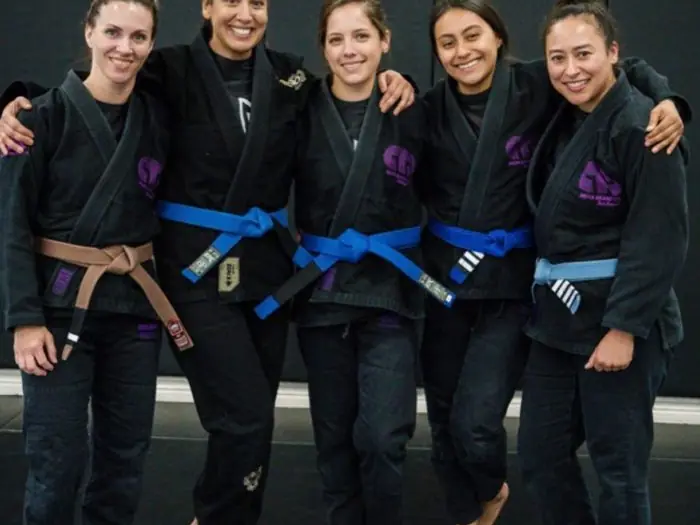 Five women in black gis with blue and brown belts pose together on the mats.